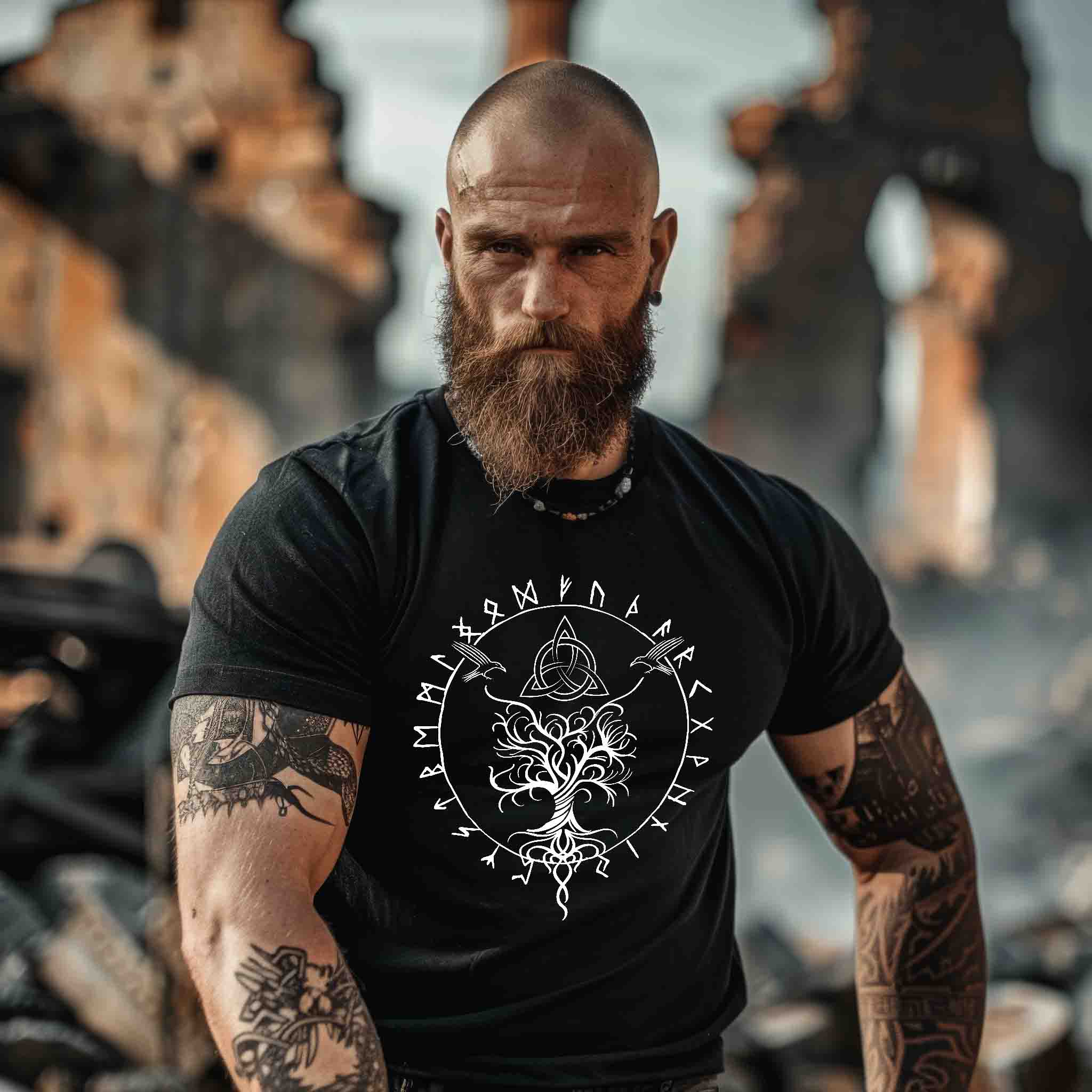 Vorderansicht eines bärtigen, tätowierten Mannes in einem schwarzen Wikinger T-Shirt mit großem weißem Frontprint von Yggdrasil, Runen und Odins Raben.“