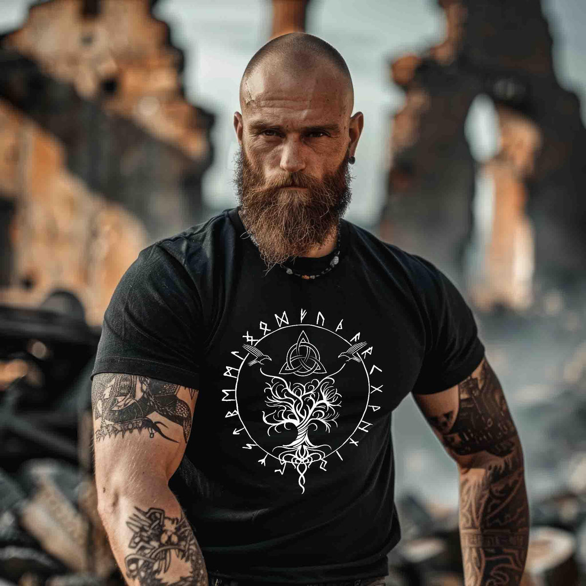 Vorderansicht eines bärtigen, tätowierten Mannes in einem schwarzen Wikinger T-Shirt mit großem weißem Frontprint von Yggdrasil, Runen und Odins Raben.“