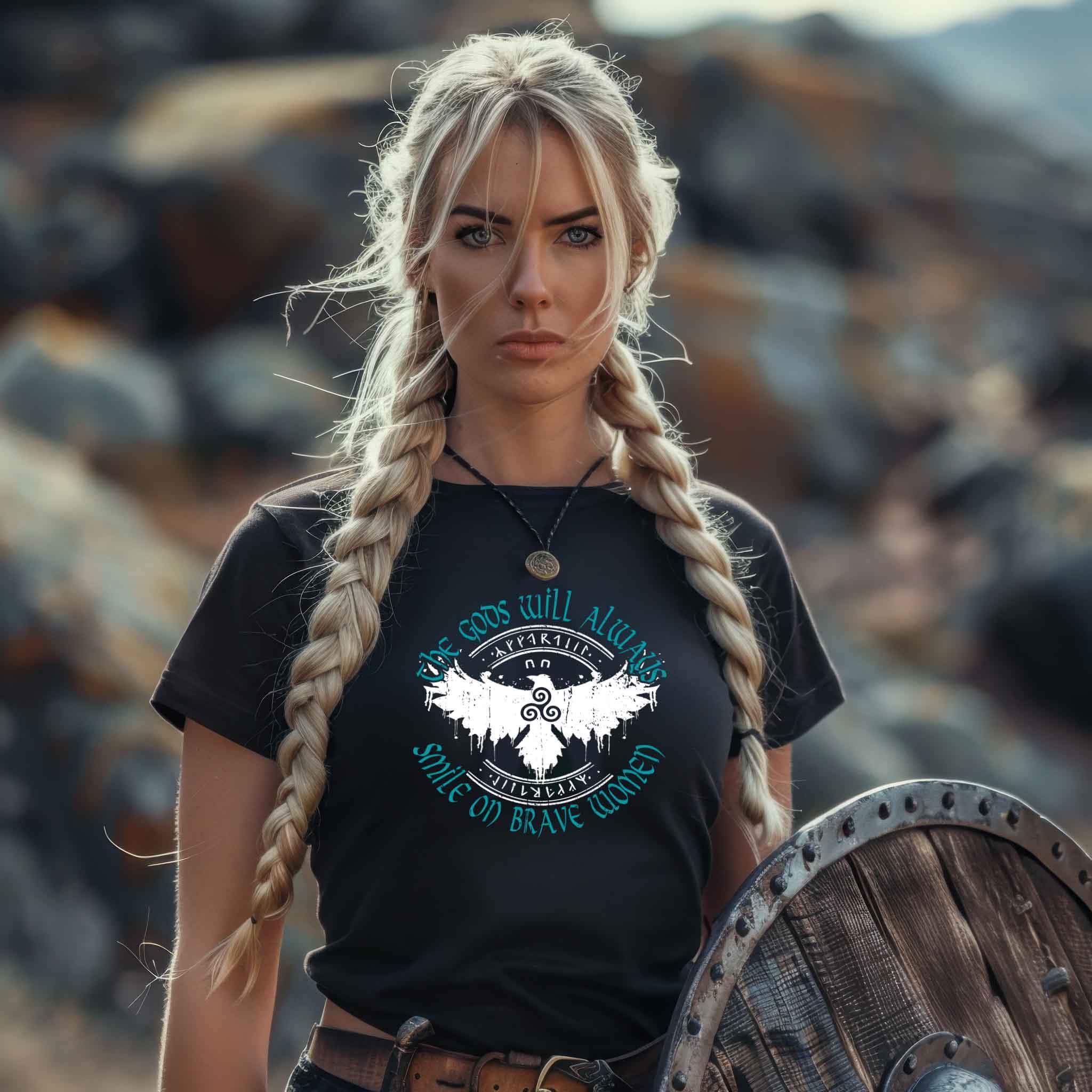 Woman wearing Schildmaiden Premiumshirt with "The Gods Will Always Smile on Brave Women" text, holding shield, outdoor setting.