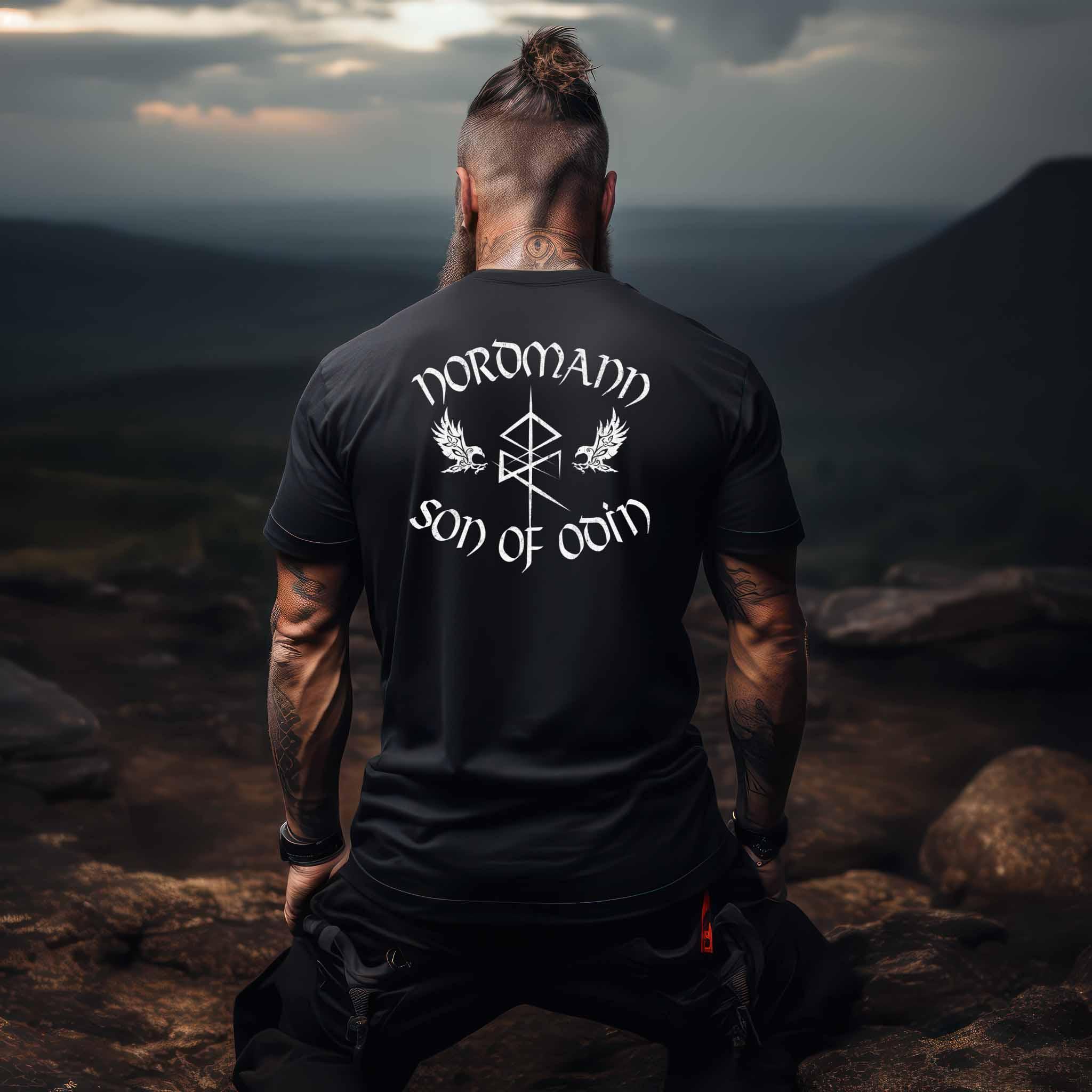 Man wearing Viking backprint shirt "Nordmann - Son of Odin" with runic symbols and ravens in a mountainous landscape.