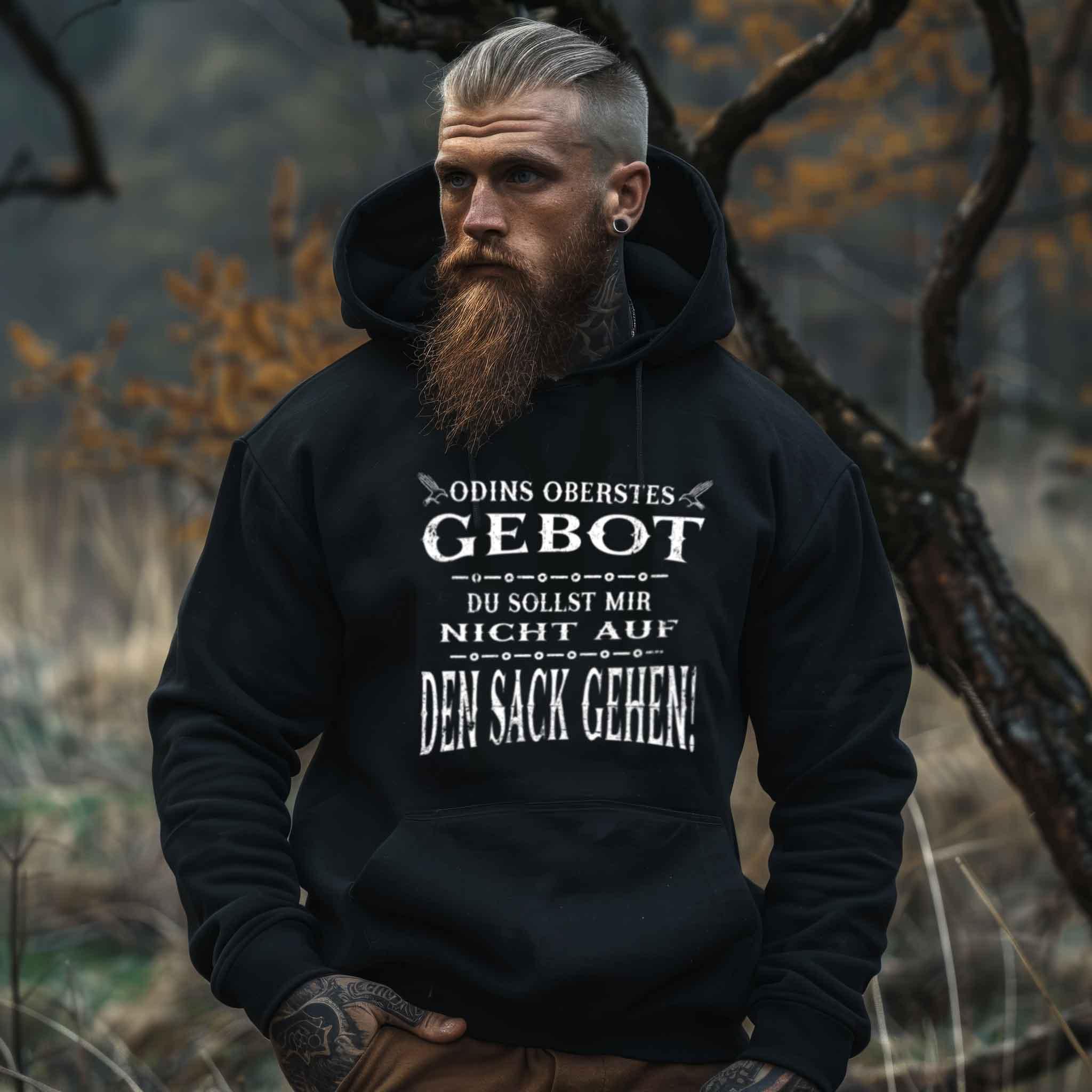 Man wearing black Viking hoodie with funny text "Odins Oberstes Gebot: Du Sollst Mir Nicht Auf Den Sack Gehen!" standing in a forest.